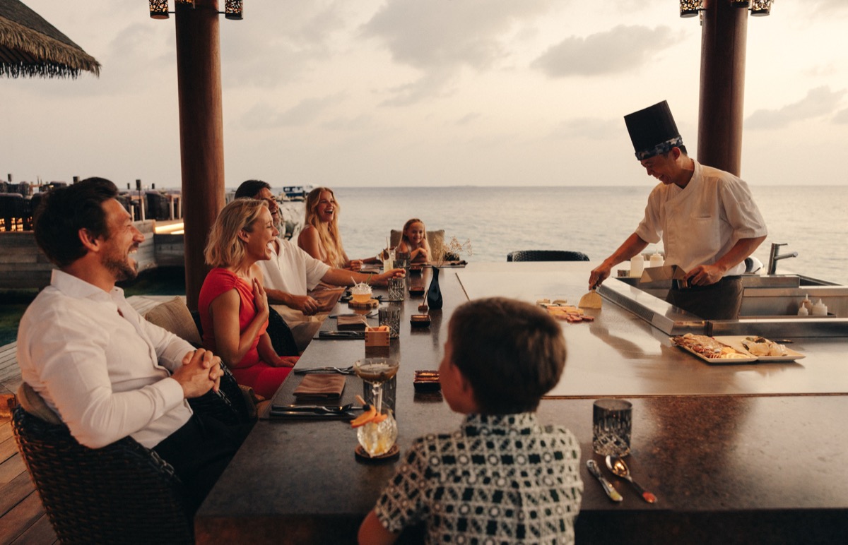 A family laughing together around a waterside restaurant counter at sunset, watching the chef work